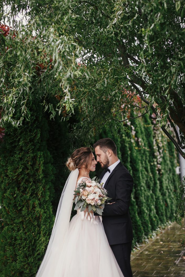 Gorgeous bride and stylish groom gently hugging on background of green trees. Sensual wedding couple embracing. Romantic moments of newlyweds. Modern wedding photo