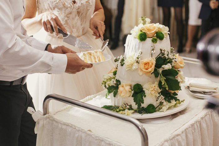 Gorgeous bride and stylish groom cutting together white wedding cake with roses at wedding reception. Happy wedding couple tasting cake. Romantic moments of newlyweds